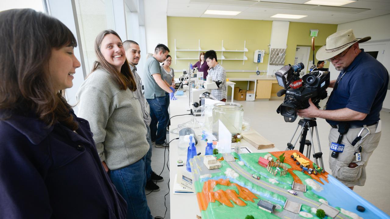 A group of AHP faculty and staff stand next to a table containing a model waterway, as a photographer sets up his camera nearby